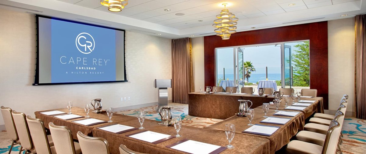 The Sandpiper meeting room at Cape Rey Carlsbad Beach set U-shape facing a screen, with an ocean-view window