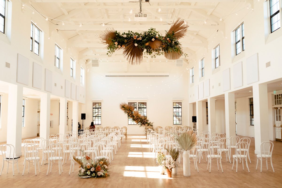 Rows of white chairs facing the front of the hall with a hanging floral installation overhead