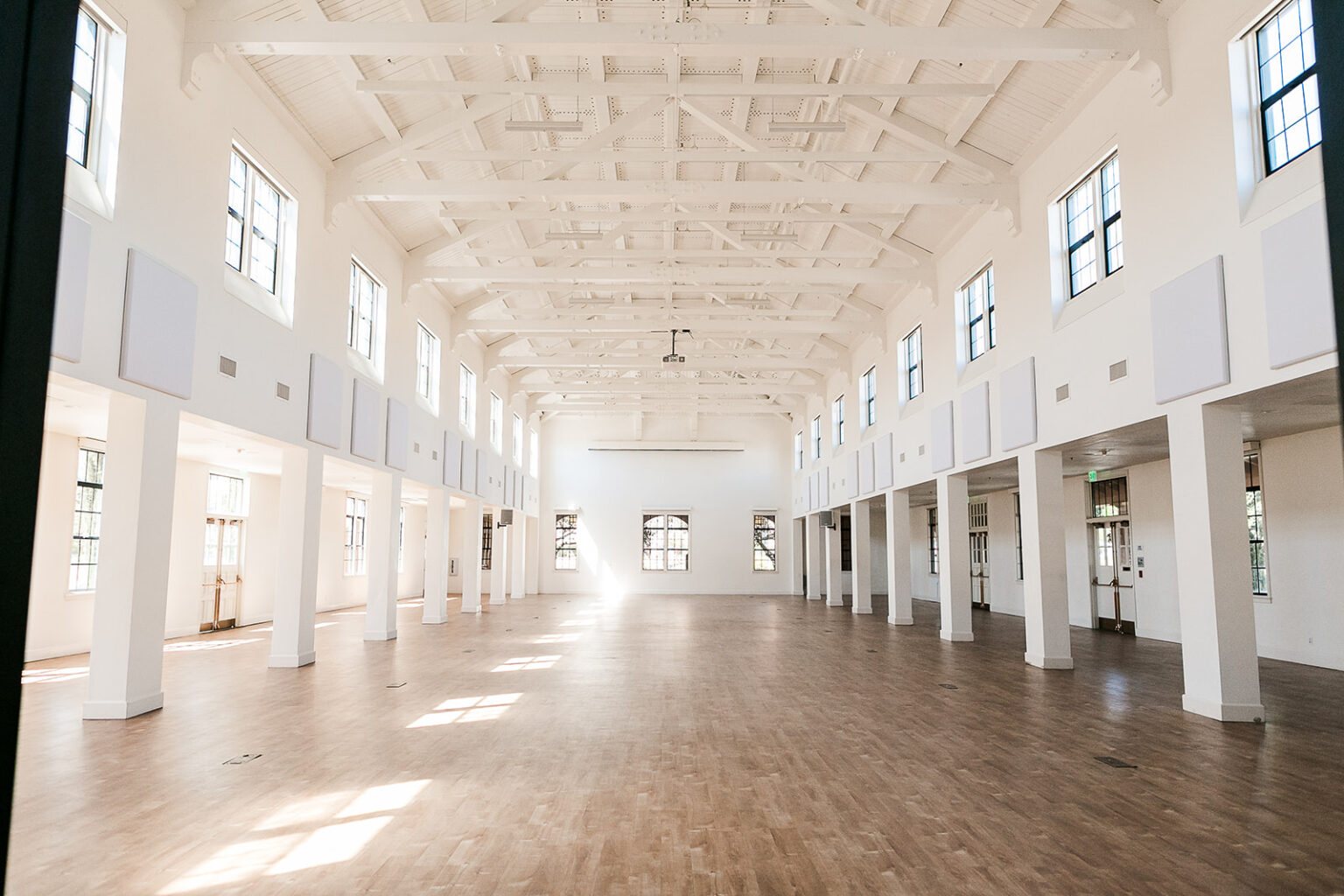 The empty Vesper Venue main hall with wood floors, white walls, tall windows and a vaulted beamed ceiling