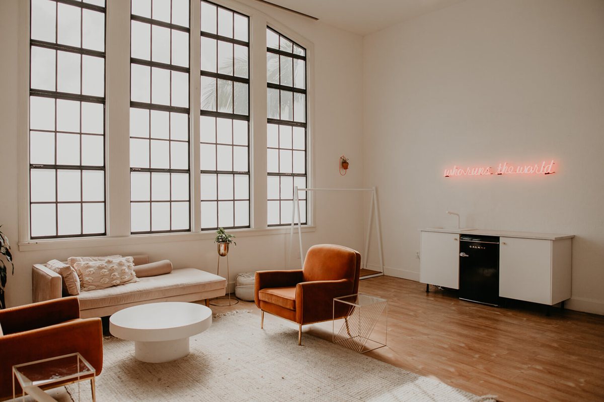 A bright private suite with tall windows, a white sofa, armchairs and a neon sign on the wall